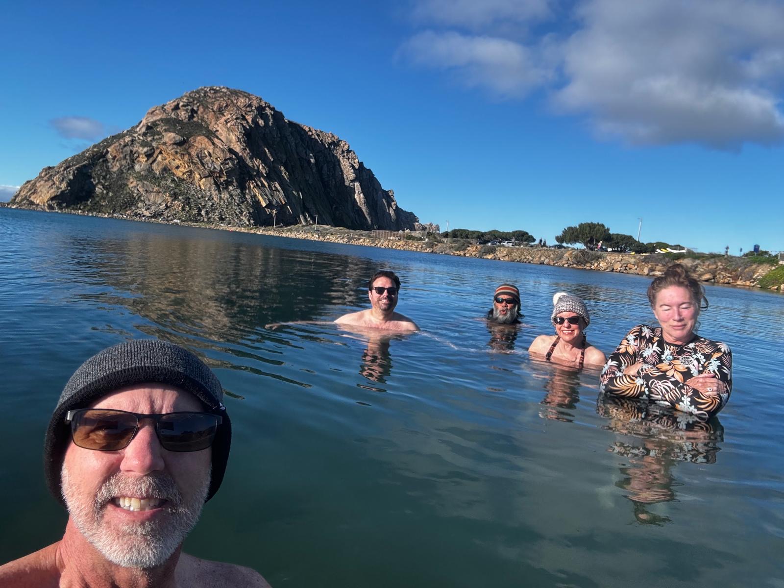 Five club members dipping together in calm water, with Russ smiling in the foreground and Morro Rock behind them